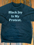 Black sweatshirt with 'Black Joy Is My Protest' text on a wooden surface