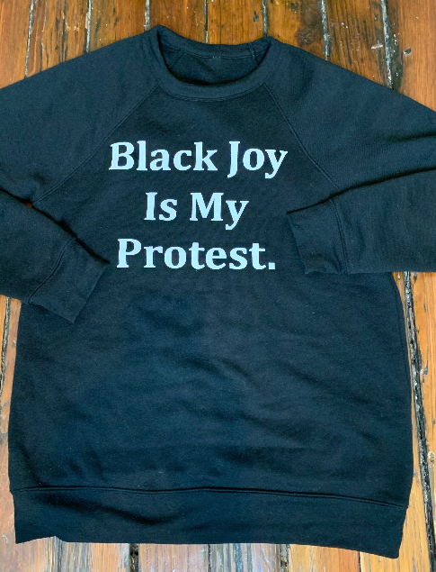 Black sweatshirt with 'Black Joy Is My Protest' text on a wooden surface